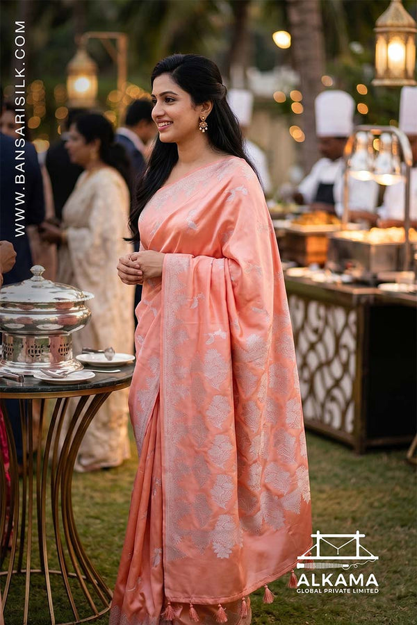 Woman in a peach-orange Banarasi Mashru silk saree at an outdoor event, with 'ALKAMA' branding.