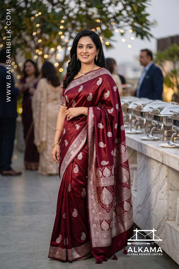 Woman in a deep red Banarasi Mashru silk saree at an outdoor event, with 'ALKAMA' branding.