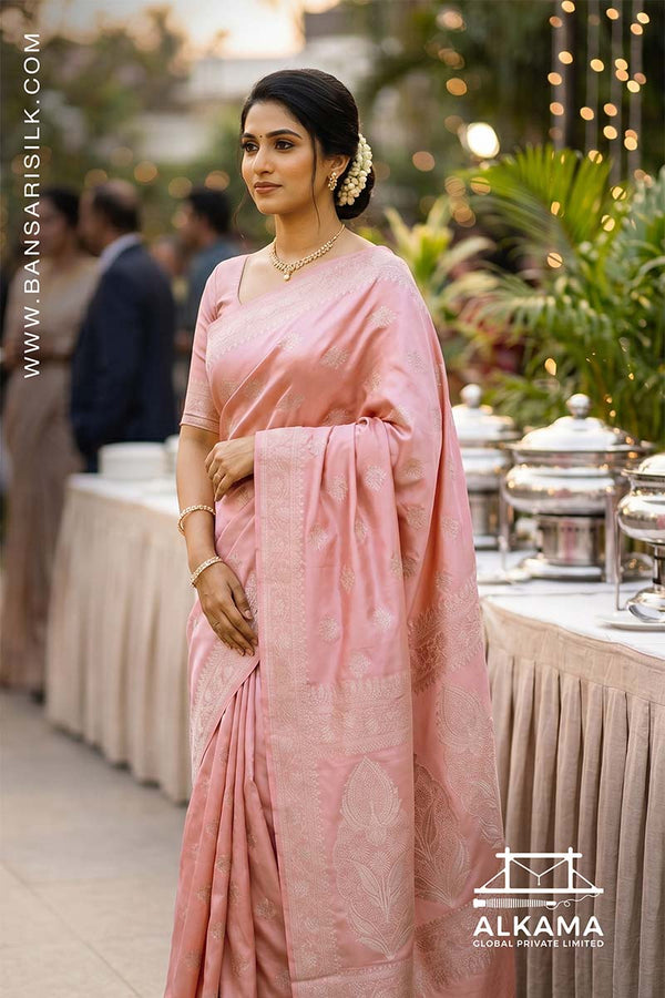 Woman in a baby pink Banarasi Mashru silk saree at an outdoor event, with 'ALKAMA' branding.