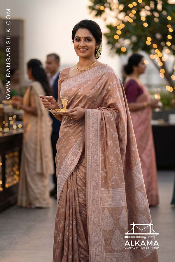 Woman in a beige-peach Banarasi Mashru silk saree at an outdoor event, with 'ALKAMA' branding.
