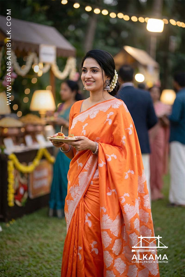 Woman in a orange Banarasi Mashru silk saree at an outdoor event, with 'ALKAMA' branding.