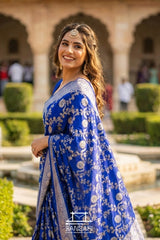 Woman in a blue Banarasi saree with floral patterns standing outdoors.