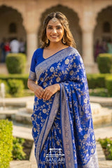 Woman in a blue Banarasi saree with floral patterns standing outdoors.