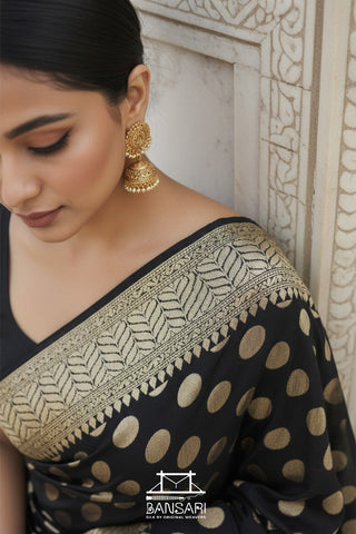 Woman wearing a black saree with gold patterns and earrings, standing against a light-colored wall.