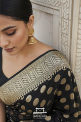 Woman wearing a black saree with gold patterns and earrings, standing against a light-colored wall.